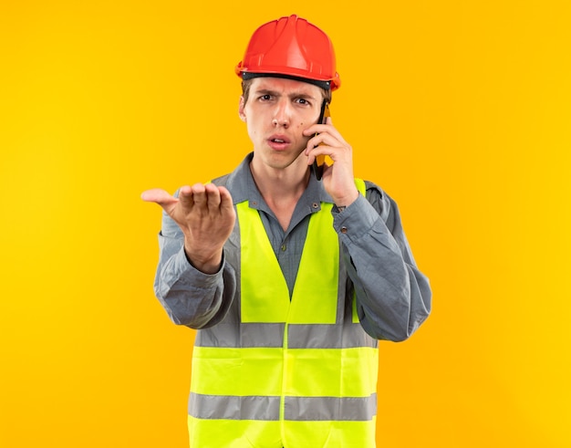 Il giovane costruttore rigoroso in uniforme parla al telefono tendendo la mano alla telecamera