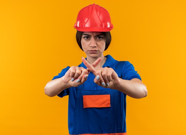 Strict young builder woman in uniform showing gesture of no isolated on yellow wall