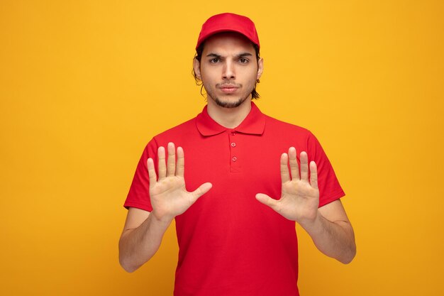 strict young delivery man wearing uniform and cap looking at camera showing stop gesture with both hands isolated on yellow background