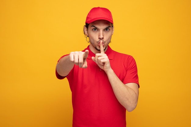 strict young delivery man wearing uniform and cap looking and pointing at camera showing silence gesture isolated on yellow background