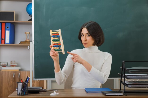 Severa giovane insegnante femminile che tiene e indica l'abaco seduto alla scrivania con gli strumenti scolastici in classe