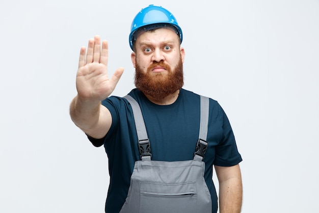 Strict young male construction worker wearing safety helmet and uniform looking at camera showing stop gesture isolated on white background