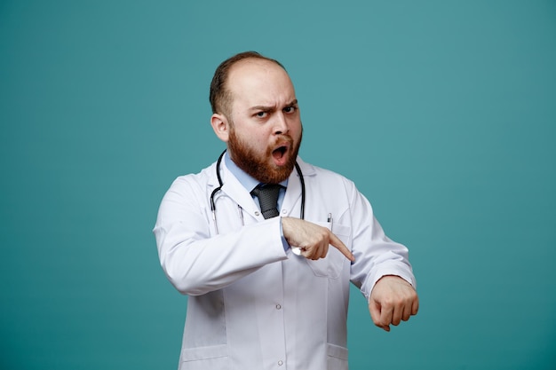 Strict young male doctor wearing medical coat and stethoscope around his neck looking at camera showing you are late gesture isolated on blue background