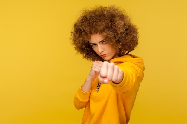 Strijd en vechtlust portret van agressieve vrouw met krullend haar in stedelijke stijl hoodie aanvallen met gebalde vuisten dreigen te slaan indoor studio shot geïsoleerd op gele achtergrond