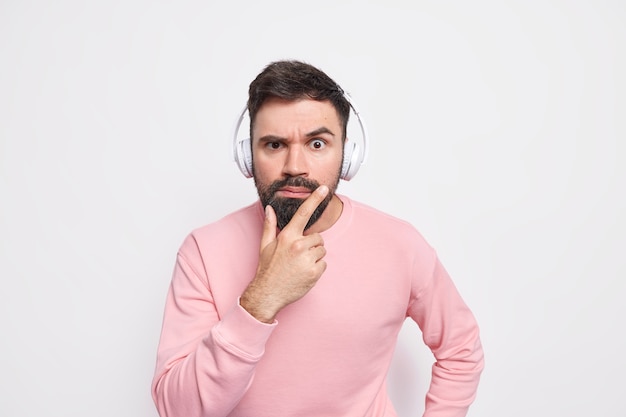 Strikt serieuze man houdt kin vast kijkt aandachtig, gefocust op iets luistert audioboek via draadloze koptelefoon gekleed in casual trui