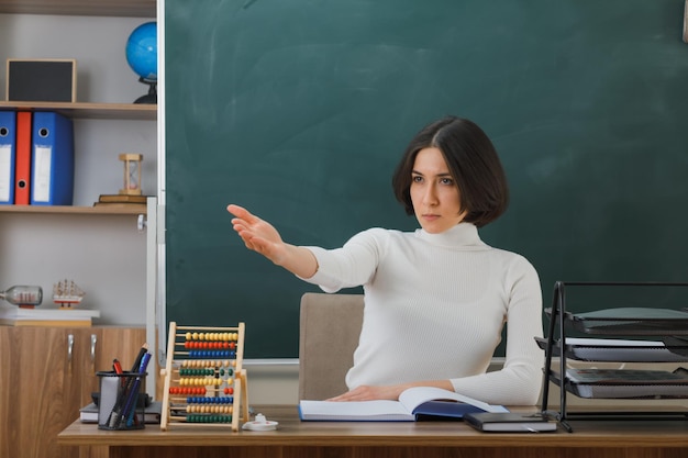 Strikte punten aan de zijkant met de hand jonge vrouwelijke leraar zittend aan een bureau met schoolhulpmiddelen in de klas