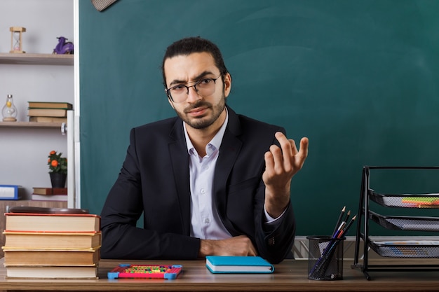 Strikte weergave kom hier gebaar mannelijke leraar met een bril die aan tafel zit met schoolhulpmiddelen in de klas