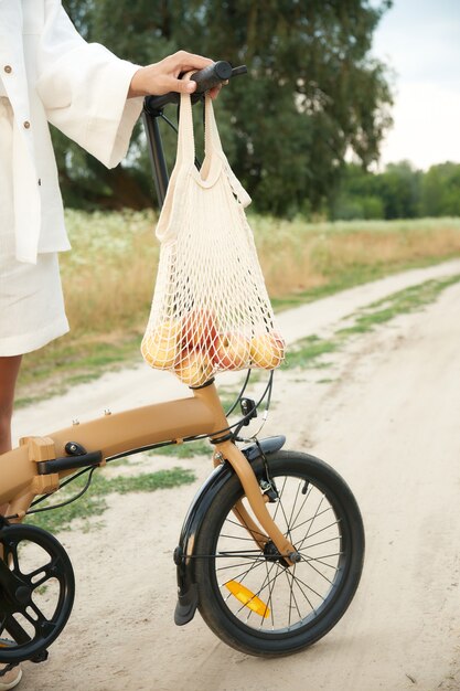 String bag with apples weighs on the handlebars of a folding bicycle next a girl in white linen suit