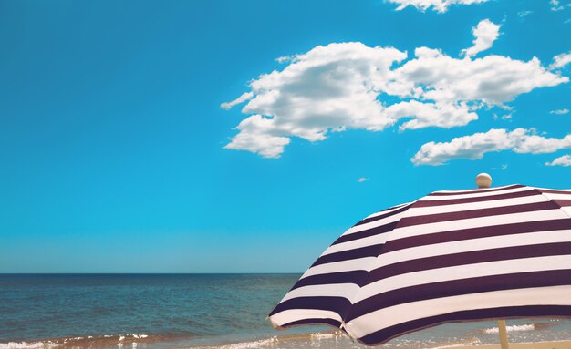 Striped  beach umbrella on the sea shore with sand, water and vivid blue sky with white clouds 