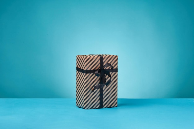 Striped brown paper present box tied with black ribbon and bow on a blue surface and background clos