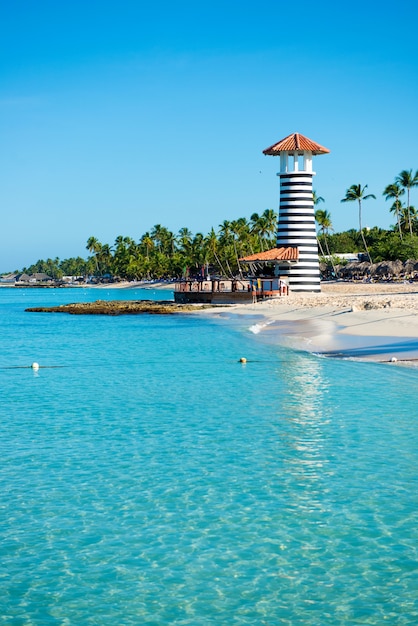 Striped lighthouse on sandy shore with palm trees. Clear water of the Caribbean sea.