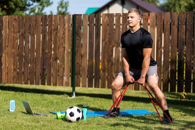 The  stromg man goes in for sports in park. Cheerful sporty man with blonde  hair  doing a squat   with rubber and watches  in laptop  in the garden