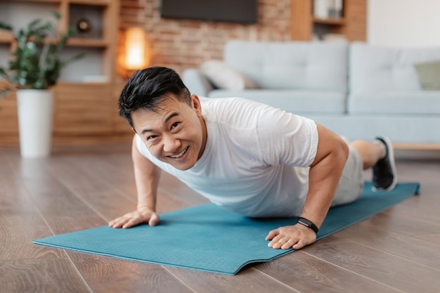Сильный азиатский зрелый мужчина делает силовые тренировки, стоя в позе доски или отжимаясь дома на коврике для йоги