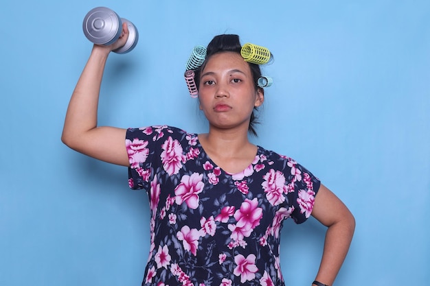 Photo strong asian woman housewife wearing home dress and hair curlers lifting dumbbel with serious expres