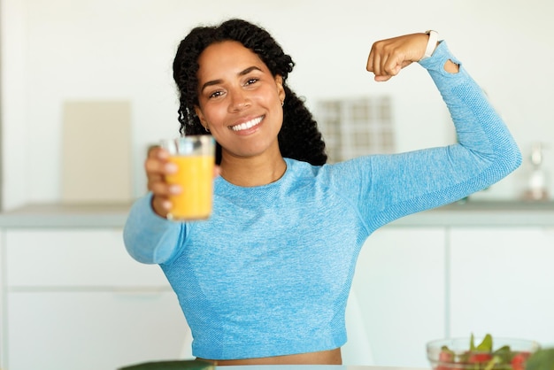 Strong athletic black woman in sportswear holding glass with juice and showing biceps standing in kitchen