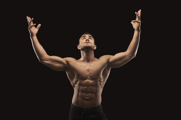 Strong athletic man showing big biceps and muscular body, standing in bodybuilder competitive posture and lookin up. Studio shot on black background. Bodybuilding concept