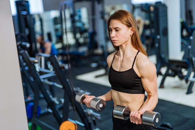 Strong athletic woman in black sport wear pumping up muscles\
with dumbbells in the gym fitness girl with perfect body in the gym\
has a training