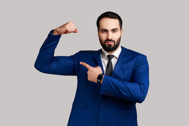 Strong bearded man raising hands showing power pointing at his biceps feeling independent strong with proud look wearing official style suit Indoor studio shot isolated on gray background