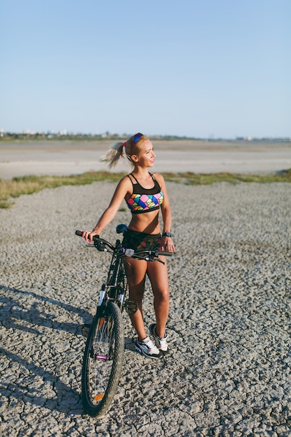 色とりどりのスーツを着た強い金髪の女性が砂漠地帯の自転車の近くに立って太陽を見ています。フィットネスのコンセプト。