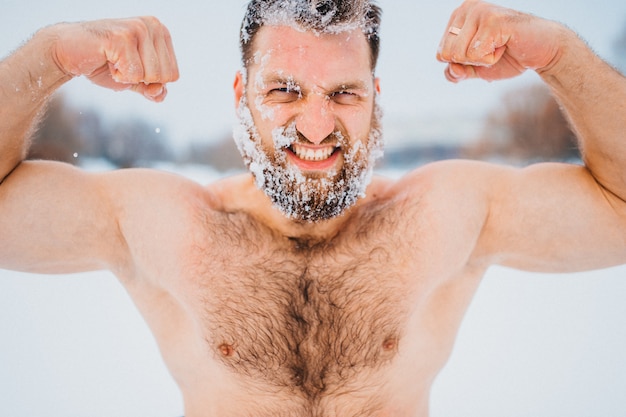 Strong bodybuilder with his face and hair covered with snow demonstrating his muscles