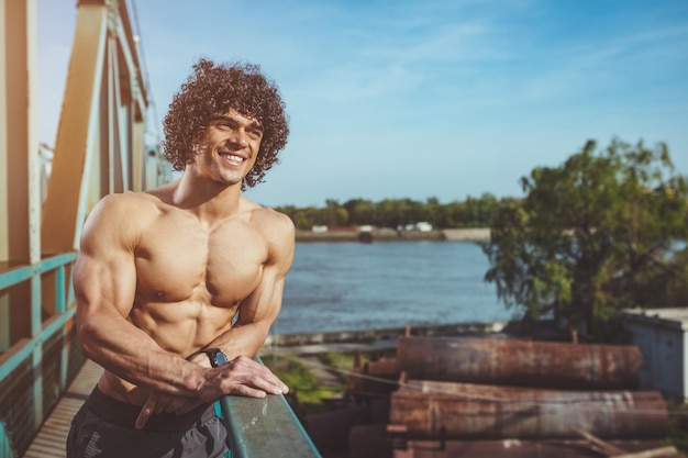 Photo strong bodybuilder with naked torso with six pack and perfect muscles standing on a bridge ready for morning workout.