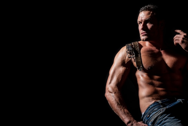 Strong brutal guy. Sexy torso. Leather belt, jeans. Handsome big muscles man posing at studio. Model showing torso with six pack abs. Isolated on black background. Showing muscular torso.