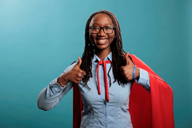 Strong and confident african american justice defender\
expressing ok symbol at camera. brave and proud superhero woman\
wearing cloak while giving thumbs up gesture sign on blue\
background.