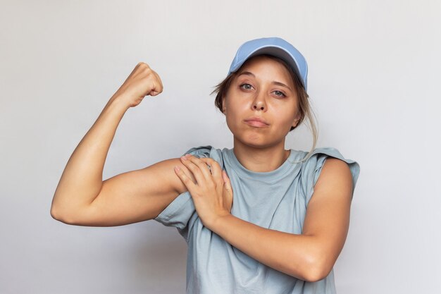 灰色のTシャツとキャップで強い自信を持って白人の若いブロンドの女性は腕を上げ、上腕二頭筋を示しています