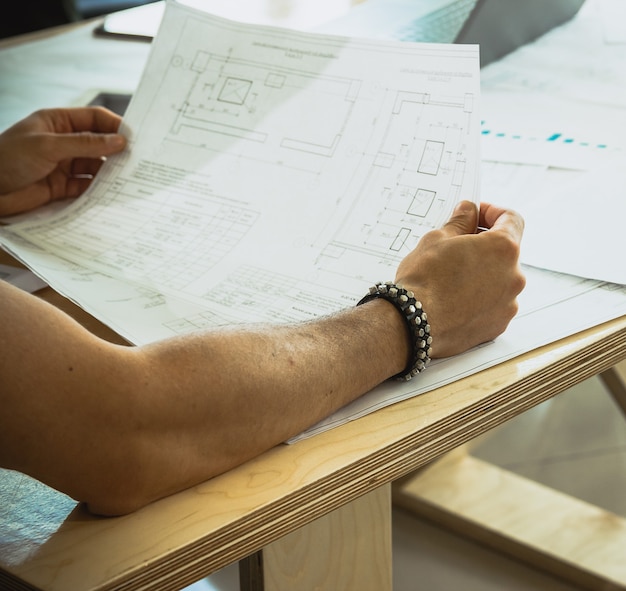 Strong courageous hands of a young man who is sitting at a wooden table holds drawings with a draft