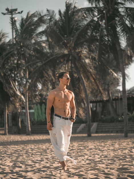 Strong fitness male model on the beach shirtless man in white\
pants