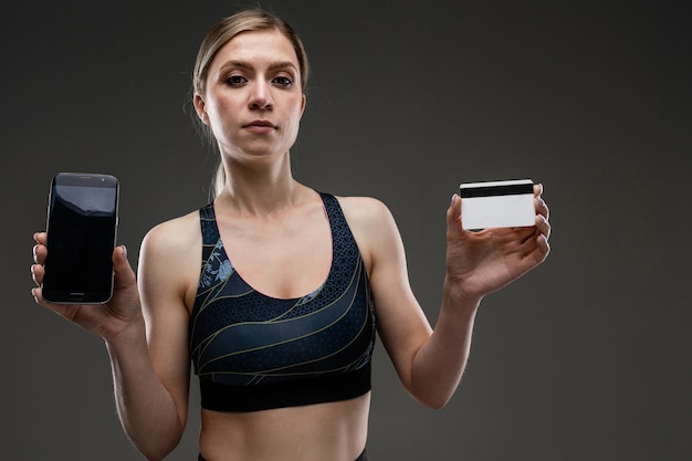 Photo strong girl in sportswear with a credit card  and a phone on a dark wall
