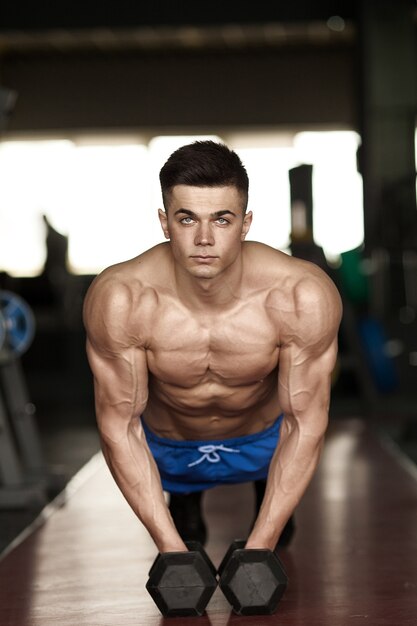 Strong, handsome man doing push-ups on dumbbells in a gym as bodybuilding exercise, training his muscles