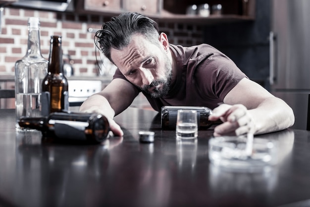 Strong hangover. Cheerless unhappy drunk man sitting at the table and waking up while feeling strong hangover