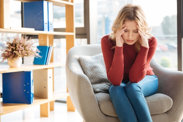 Strong headache. Unhappy cheerless depressed woman sitting in the armchair and holding her temples while having a strong headache