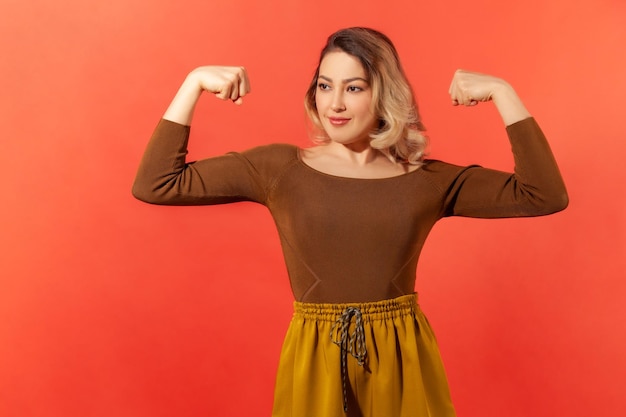 Strong independent woman with blond hair raised arms and showing her muscles proud of her strength and leadership skills emancipation Indoor studio shot isolated on red background