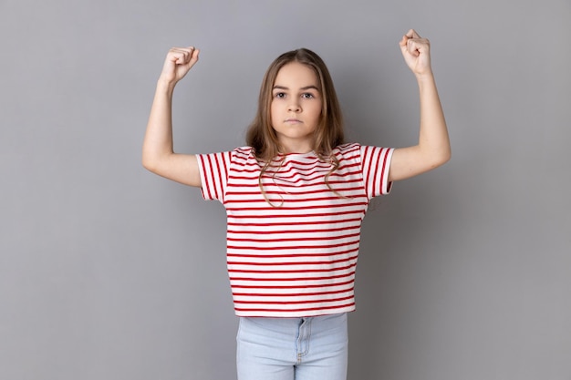 Photo strong little girl standing and raising arms showing her strength and looking at camera