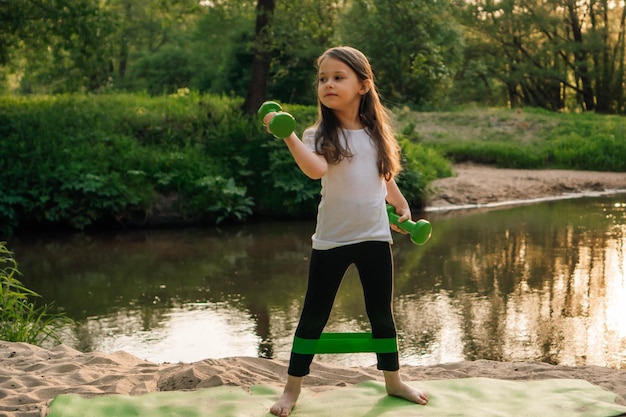 Strong little girl with mini band on legs working out on beach in front of lake standing on mat and lifting dumbbells