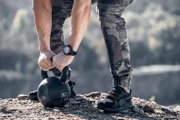 Strong male arms on the kettlebell of 24 kilos Muscular hands with a watch on the weight Bodybuilders hands and legs close up on the rock