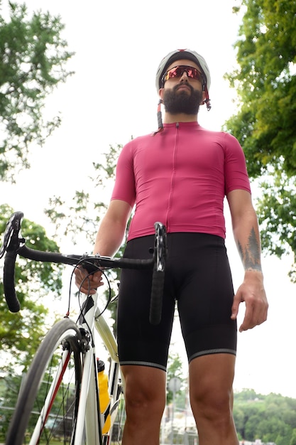 Foto ciclista maschio forte con occhiali sportivi e casco protettivo che cammina con la sua bicicletta nella foresta per fare una pausa dopo aver guidato sky blue e foresta sullo sfondo