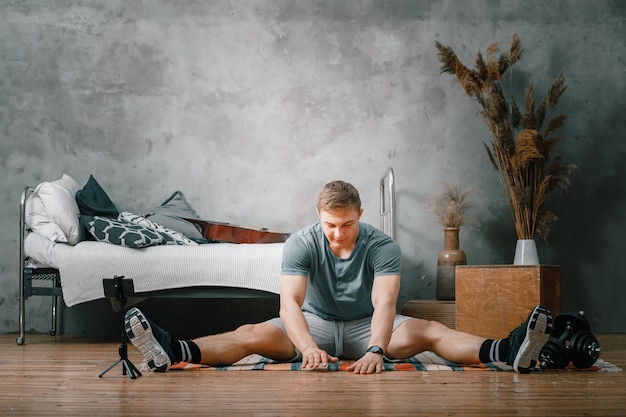 The strong man goes in for sports at home. Cheerful sporty man with blond  hair stretches to the leg  and shooting  blog  in the bedroom