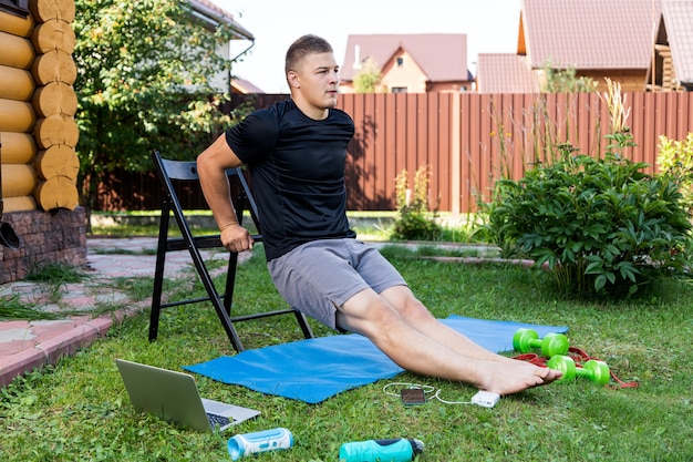 The  strong man goes in for sports in park. Cheerful sporty man with black  hair  doing a squat   with chair and watches  in laptop  in the garden