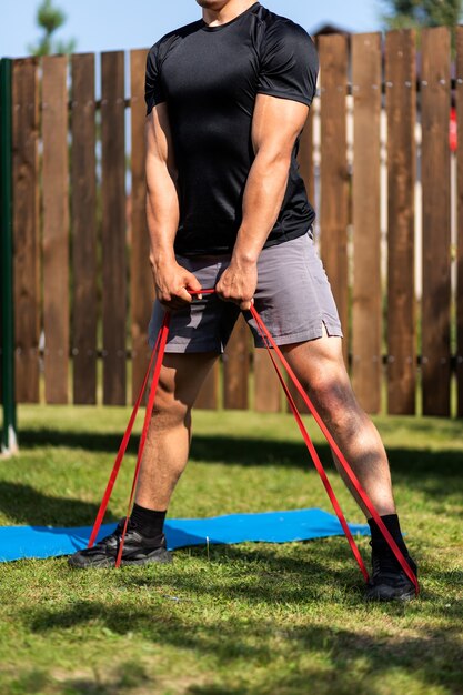 The  strong man goes in for sports in park. Cheerful sporty man with blonde  hair  doing a squat   with rubber and watches  in laptop  in the garden