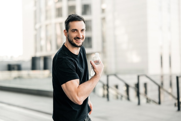 Strong man shows biceps, holds plastic water bottle, looks at camera and smiles. Morning city on background. Attractive man excercised a lot to get a fit body. Hydration time!