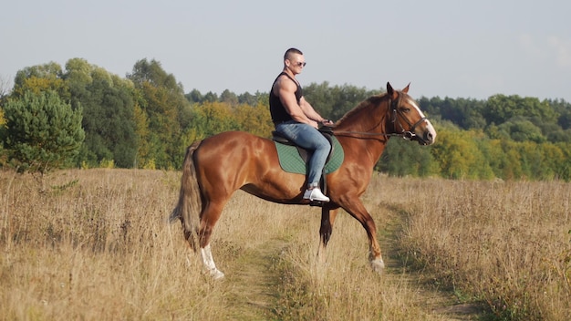 A strong man in a tshirt of black color in denim pants and sunglasses riding a horse in nature