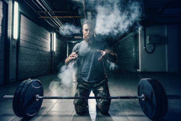 Strong muscular Caucasian bodybuilder clapping hands and crouching in hall. In front of him barbell, chalk all around. Back light, night workout concept.