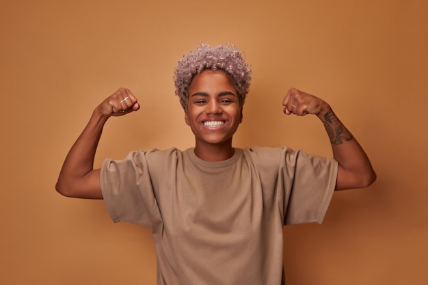 Photo strong powerful african american woman with blonde curly hair toothy smile raises arms and shows bic...