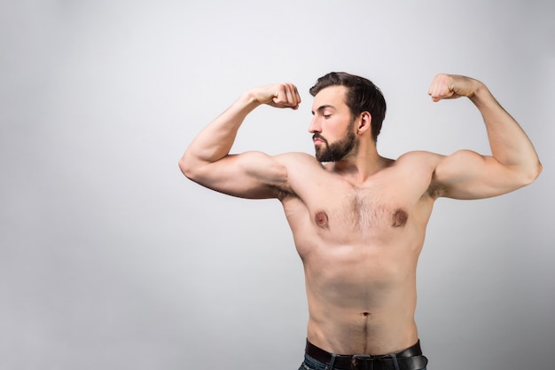 Ragazzo forte e potente senza camicia è in piedi al muro bianco e in posa. sta mostrando i suoi grandi muscoli e quanta forza ha. ama se stesso. taglia vista.