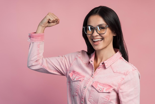 腕の筋肉が手を上げて、自由で独立していることを示す強い自信のあるブルネットの女性。解放の概念。ピンクの背景に分離された屋内スタジオショット