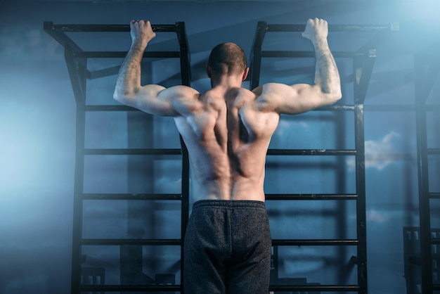 Strong sportsman training on horizontal bar in gym, back view.