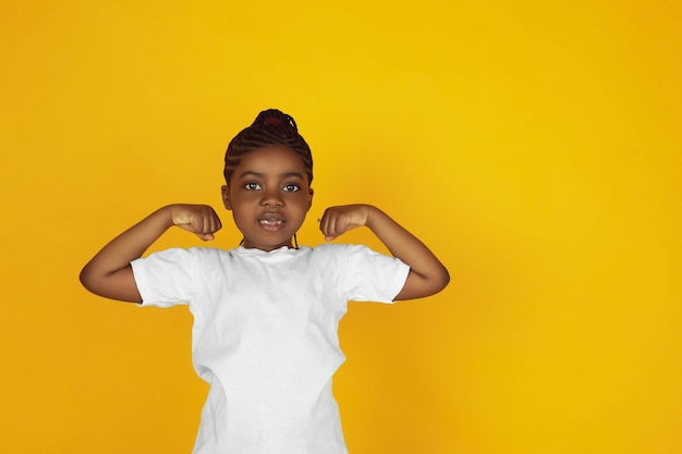 강한, 승자, 리더. 노란색 스튜디오 배경에 있는 작은 아프리카계 미국인 소녀의 초상화. 명랑하고 아름다운 아이. 인간의 감정, 표현, 판매, 광고의 개념. 카피스페이스. 귀여워 보인다.
